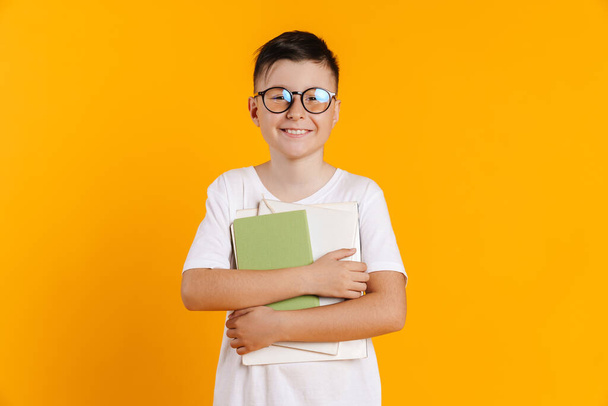 Brunette white boy in eyeglasses smiling while posing with exercise books isolated over yellow background - Фото, изображение