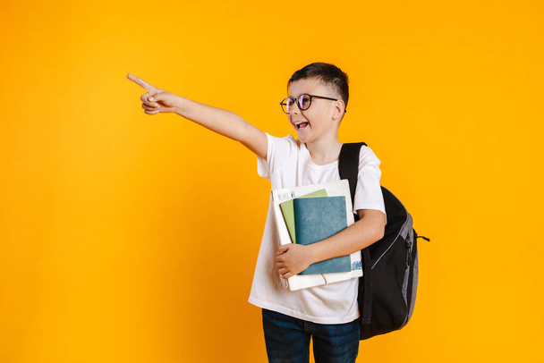 Happy smiling preteen boy in t-shirt standing over yellow background wearing backpack holding textbooks pointing finger aside - Photo, image