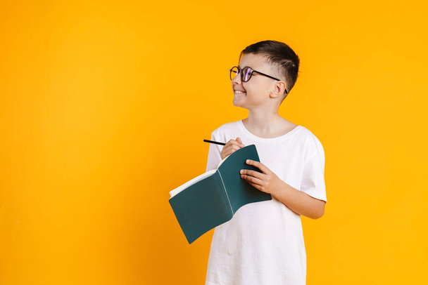 Happy smiling preteen boy in t-shirt standing over yellow background taking notes in a diary - Fotoğraf, Görsel