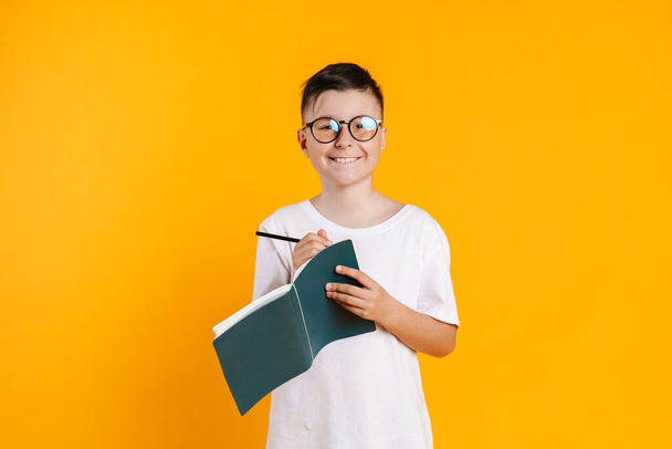 Happy smiling preteen boy in t-shirt standing over yellow background taking notes in a diary - Valokuva, kuva