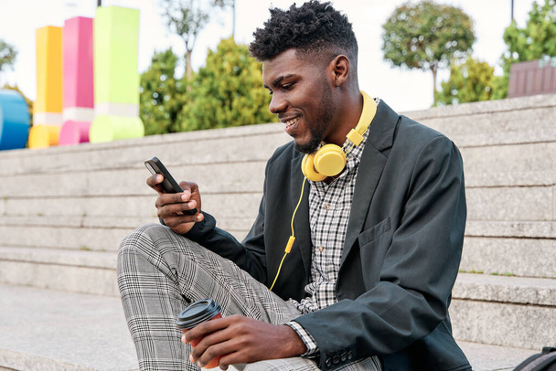 Jovem africano alegre vestindo blazer ao ar livre, segurando taça de café takeaway, usando telefone celular - Foto, Imagem