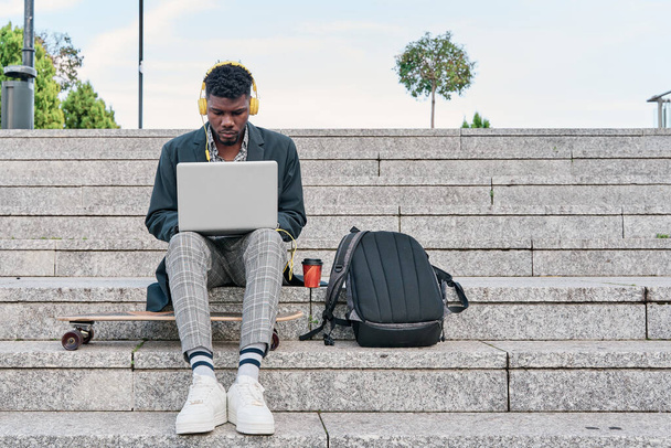 front view of an african american man sitting on a staircase working on his laptop. hipster man listening to music with yellow headphones. - Foto, immagini