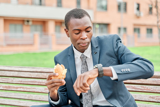 Afroamerikanischer Geschäftsmann isst einen Hamburger, sieht Armbanduhr auf einer Bank im Park sitzen. - Foto, Bild