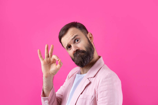 Portrait of a smiling young man in a pink jacket. Man showing ok gesture while standing in studio over isolated red background. - Fotoğraf, Görsel
