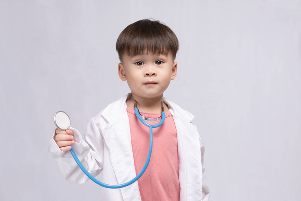 Joven chico asiático lindo usando un uniforme médico sosteniendo un estetoscopio jugando médico feliz sobre fondo blanco. Niños preescolares pretenden ser pediatras. - Foto, Imagen
