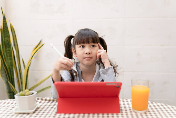 Aziatisch meisje studeren online op haar bureau thuis met tablet en pen. Thuisonderwijs tijdens de quarantaine. Blijf thuis entertainment. kinderen en tablet, Kinderen denken en analyseren van huiswerk. - Foto, afbeelding