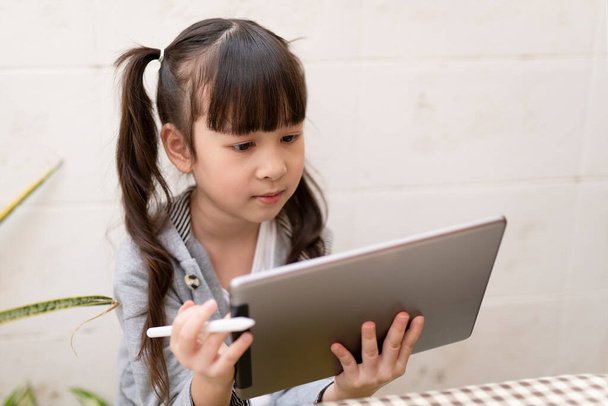 Asian girl with digital tablet and pen studying online at home. Education and distance learning for kids. Homeschooling during quarantine. Stay at home entertainment. - 写真・画像