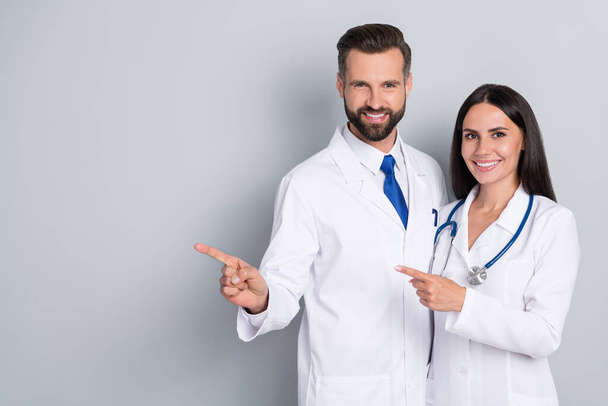 Photo of positive pretty two doctors wear white coats pointing empty space hugging isolated grey color background - Foto, imagen