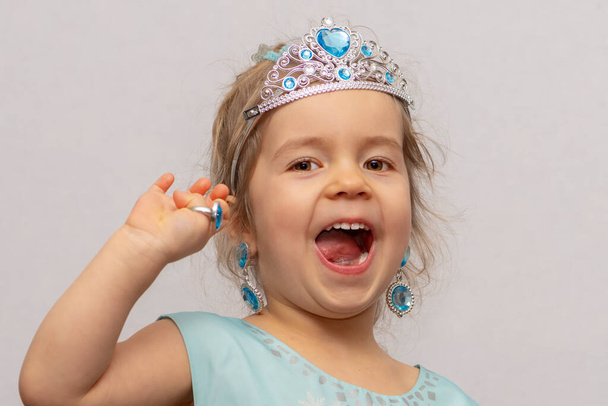 Pieni tyttö kruunussa ja korvakoruissa osoittaa lelurenkaita sormillaan, lasten koruilla ja pukukoruilla.. - Valokuva, kuva