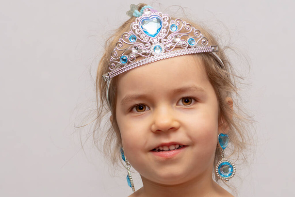 A little girl in a crown and earrings demonstrates toy rings on her fingers, children's jewelry and costume jewelry. - Foto, Imagen