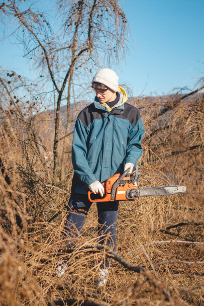 young 17-year-old temporary worker in work clothes wrestles with a chainsaw and a dry larch tree. Wood processing in the wilderness. Cutting a fallen tree into parts. Learning a new workforce.  - Photo, Image