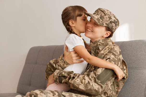 Horizontal shot of satisfied little girl with m other, military woman wearing camouflage uniform and cap posing with her daughter, family hugging each other and expressing happiness. - Photo, image