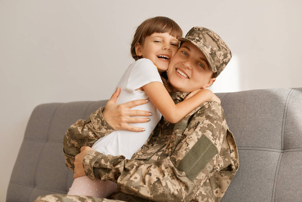 Indoor shot of delighted extremely happy military woman wearing camouflage uniform and cap posing with her daughter at home while sitting on sofa, hugging, mother come back home from war. - Photo, image
