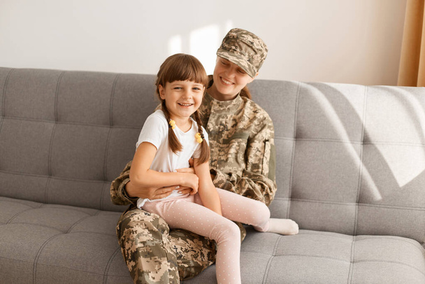 Horizontal shot of happy Caucasian woman wearing camouflage uniform and cap posing with her daughter, sitting on sofa, expressing positive emotions and happiness. - Fotografie, Obrázek