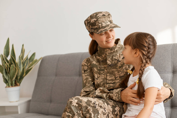 Indoor shot of smiling soldier female sitting and looking at her kid with smile, being happy to return home from army, woman wearing camouflage uniform and cap posing with her daughter - 写真・画像