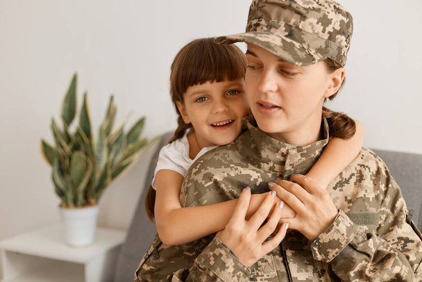 Portrait of happy positive Caucasian woman wearing camouflage uniform and cap posing with her daughter, little cute hugging her mother, missing so much. - Valokuva, kuva