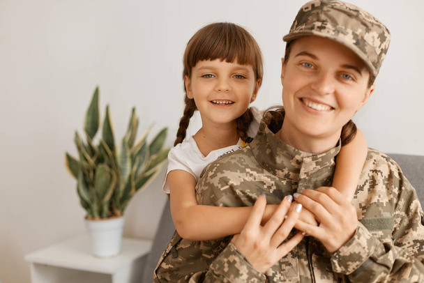 Portrait of Caucasian smiling mother and her child, woman wearing camouflage uniform and cap posing with her daughter, looking at camera with happy toothy smile. - Photo, image