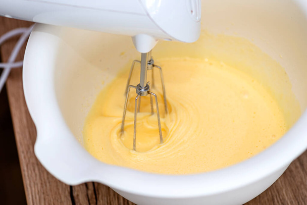 Kneading ciasto za pomocą miksera ręcznego z trzepaczką, ciasto jest w żółtym stanie ciekłym. - Zdjęcie, obraz