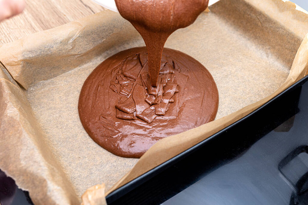 Наливаючи рідку товсту масу тіста з какао на лист для випічки, вистелений папером для випічки
. - Фото, зображення