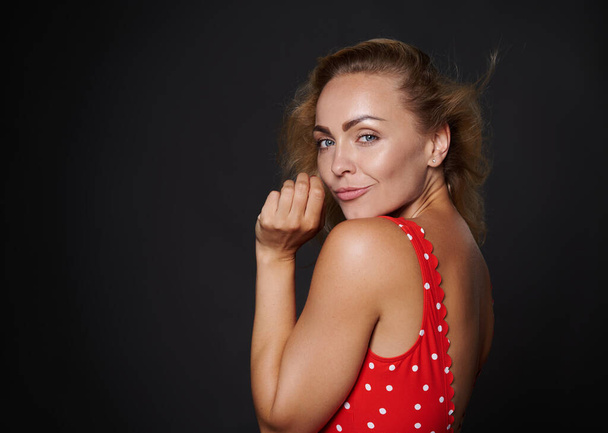 Aantrekkelijke Europese vrouw met gezonde schone frisse gloeiende gebruinde huid en blond glanzend recht haar, natuurlijke make-up, het dragen van rode badpak met witte stippen geïsoleerd op zwarte achtergrond - Foto, afbeelding