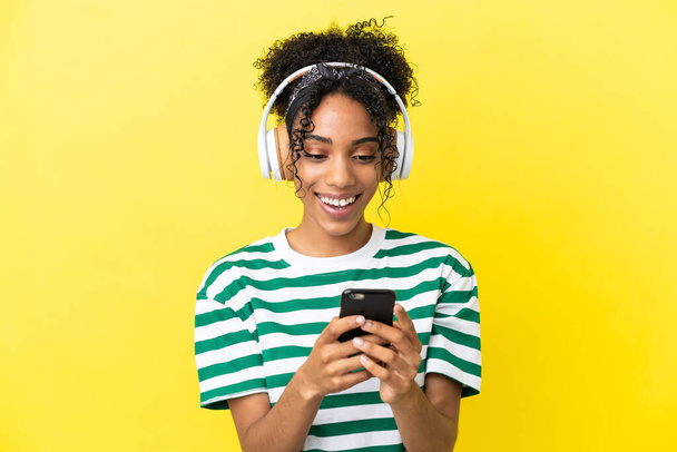 若いアフリカ系アメリカ人の女性は黄色の背景に音楽を聞いて孤立し、モバイルを探しています - 写真・画像