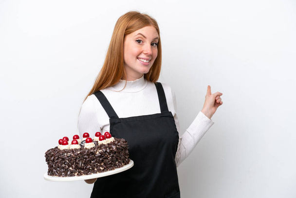 Молодая рыжая женщина держит торт на белом фоне, указывая назад - Фото, изображение