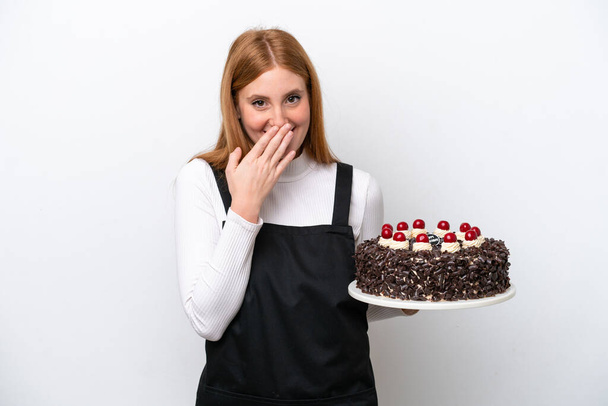 jonge roodharige vrouw houden verjaardag taart geïsoleerd op witte achtergrond gelukkig en glimlachende bedekking mond met de hand - Foto, afbeelding