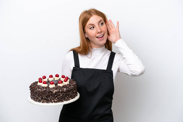 Jeune rousse femme tenant gâteau d'anniversaire isolé sur fond blanc écoutant quelque chose en mettant la main sur l'oreille - Photo, image