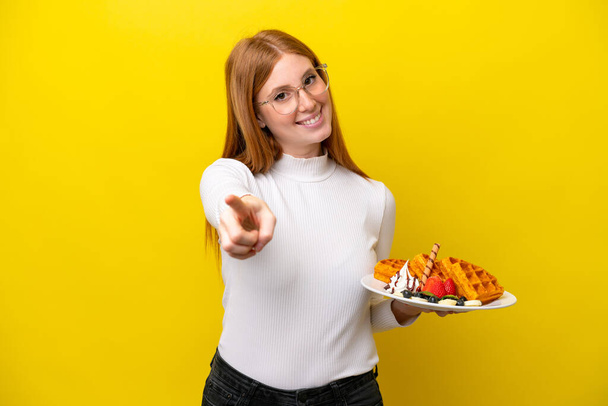 Young redhead woman holding waffles isolated on yellow background pointing front with happy expression - Foto, imagen