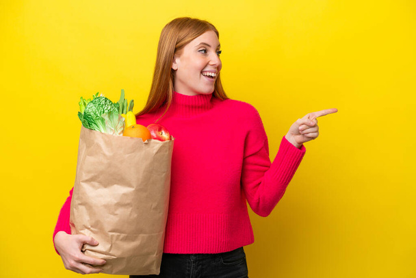 Young redhead woman holding a grocery shopping bag isolated on yellow background pointing finger to the side and presenting a product - Photo, Image