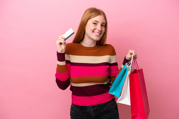 Joven pelirroja aislada sobre fondo rosa sosteniendo bolsas de compras y una tarjeta de crédito - Foto, imagen