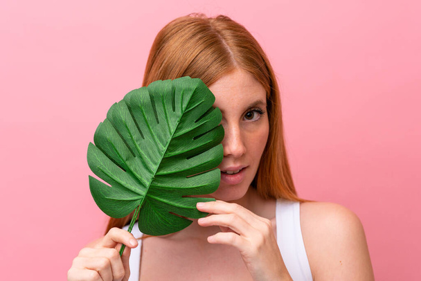 Young redhead woman isolated on pink background holding a palm leaf. Close up portrait - Fotografie, Obrázek