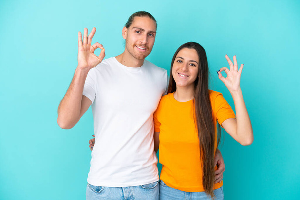 Νεαρό καυκάσιο ζευγάρι απομονώνονται σε μπλε φόντο δείχνει μια πινακίδα εντάξει με τα δάχτυλα - Φωτογραφία, εικόνα
