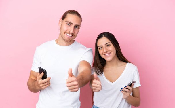 Young caucasian couple isolated on pink background using mobile phone while doing thumbs up - Photo, Image