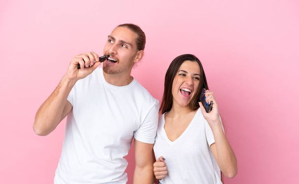 Jeune couple caucasien isolé sur fond rose en utilisant le téléphone mobile et le chant - Photo, image