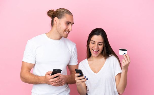 Joven pareja caucásica aislada sobre fondo rosa comprando con el móvil con una tarjeta de crédito - Foto, Imagen