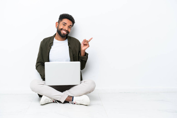 Young Brazilian man with a laptop sitting on the floor isolated on white pointing finger to the side - Fotoğraf, Görsel