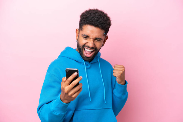 Young Brazilian man isolated on pink background using mobile phone and doing victory gesture - 写真・画像