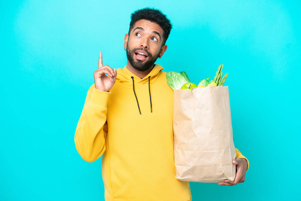 Young Brazilian man taking a bag of takeaway food isolated on blue background thinking an idea pointing the finger up - Foto, Imagen
