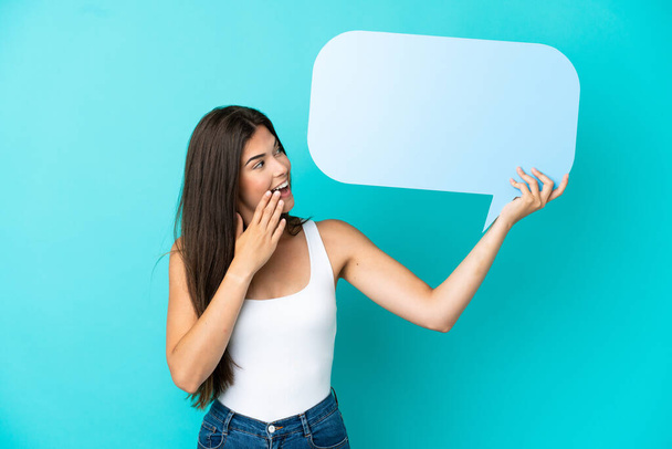 Young Brazilian woman isolated on blue background holding an empty speech bubble with surprised expression - Фото, изображение