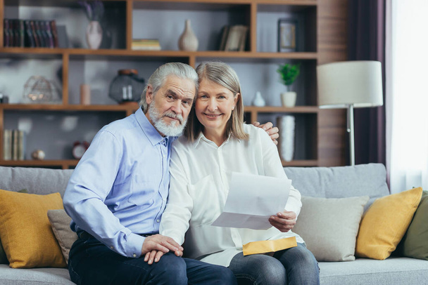 Senior couple man and woman together at home, received good news letter, happy and smiling sitting on sofa, looking at camera - Fotoğraf, Görsel