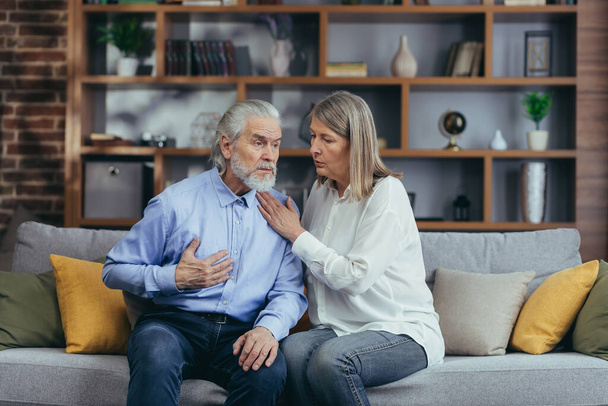 Senior mature woman helping her husband with chest pain. Old man feeling bad pain disease. Wife supporting husband - Elderly couple suffering from symptoms heart attack ache. sitting at home indoors - Photo, image