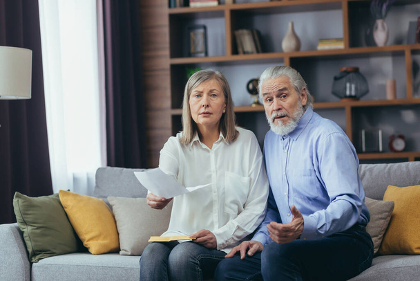 Frustrated married couple, retired man and woman, sitting on sofa at home, frustrated with document and letter, looking at camera - Фото, изображение