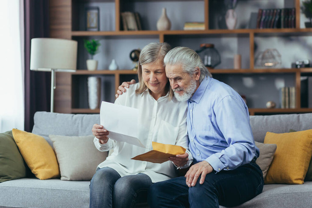 Happy senior retired couple sitting on sofa at home reading letter, happy and smiling - Photo, image