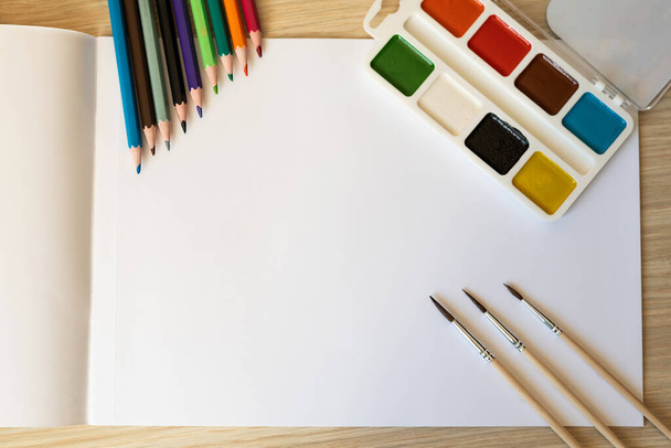 A white sheet for drawing, watercolors and brushes, pencils lie on a white table. Background for writing, school theme. - Photo, Image
