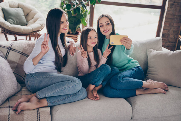 Photo of cheerful ladies recording video smartphone show fingers peace v-symbol sit divan apartment indoors - Foto, imagen