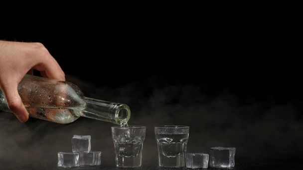 Bartender pouring up two shots of vodka with ice cubes from bottle into glasses on black background - Photo, Image