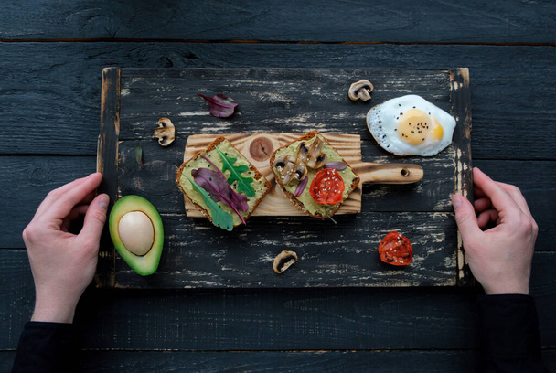 Hands holding board met avocado toast met gegrilde tomaten, gegrilde champignons, gebakken ei en salade op donkere houten tafel. Gezonde voeding en veganistische dieetconcepten - Foto, afbeelding