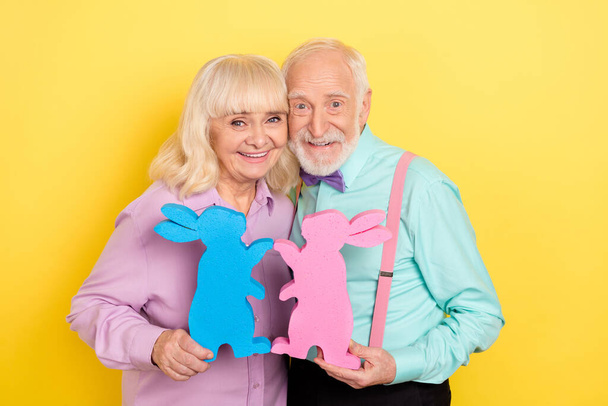 Portrait of two attractive trendy cheery grey-haired spouses holding festal bunny isolated over bright yellow color background - Foto, Imagen