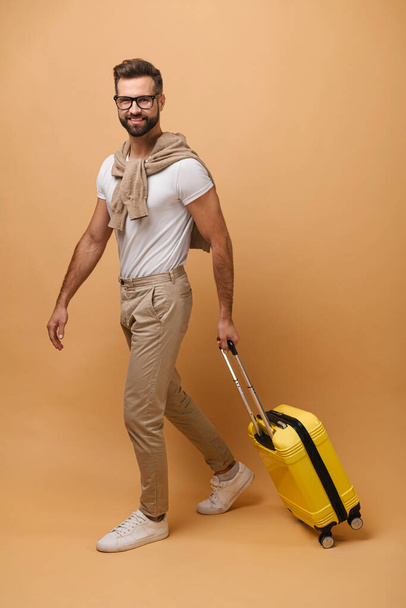 Full length view of the smiling positive man carrying luggage and stepping somewhere while looking at the camera. Traveling - Foto, imagen